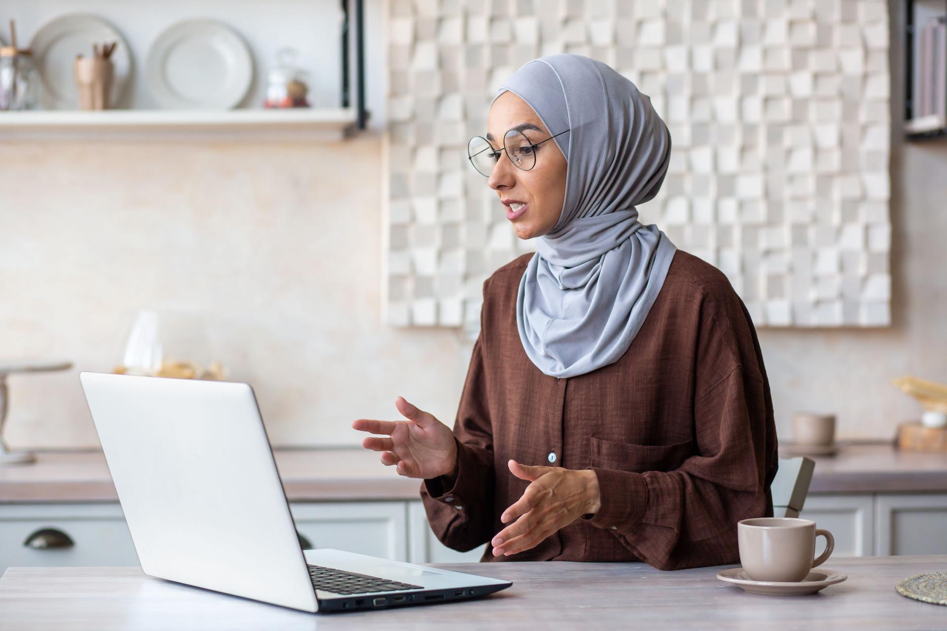 Online psychotherapy session. A young female Muslim psychologist in a hijab sits at home in front of a laptop and conducts a remote therapy consultation with a patient